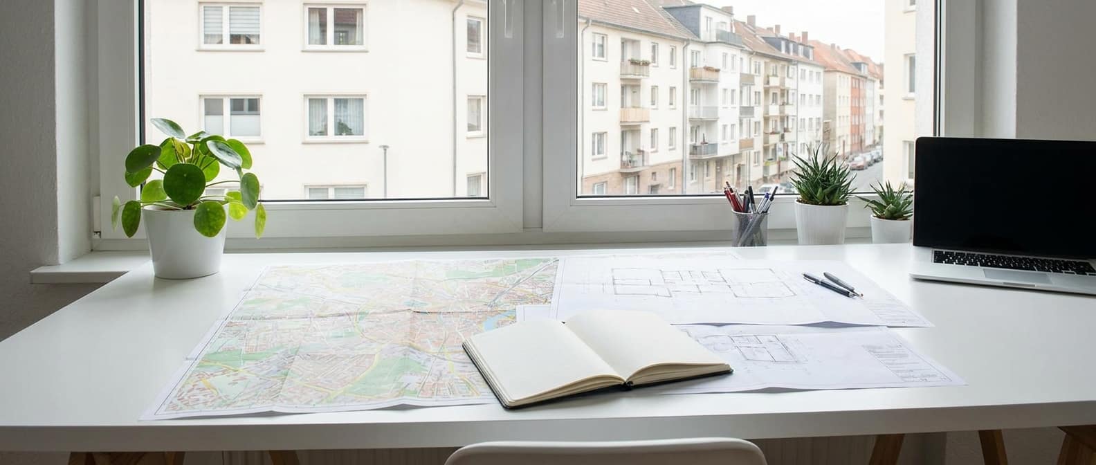 Heller Schreibtisch mit Stadtplan und Bauplan am Fenster mit Blick auf Wohnhäuser